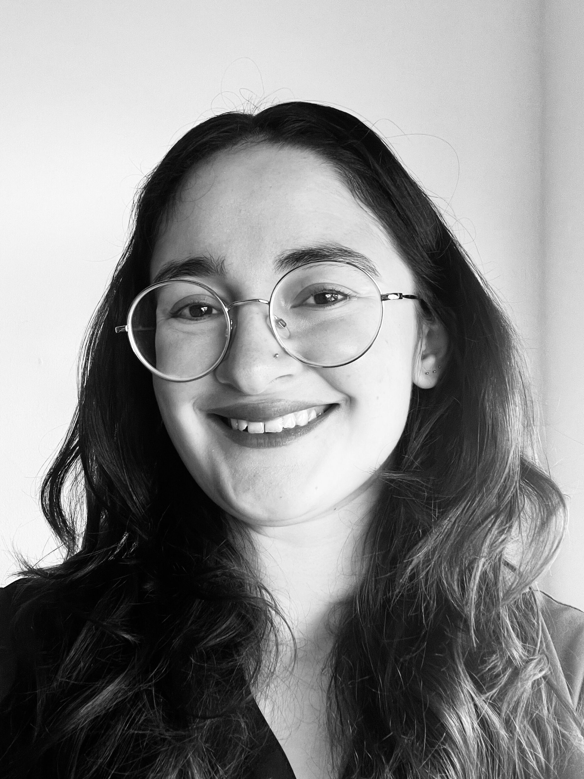 Smiling person with long, wavy hair wearing glasses and a dark top, looking directly at the camera in a black and white portrait.