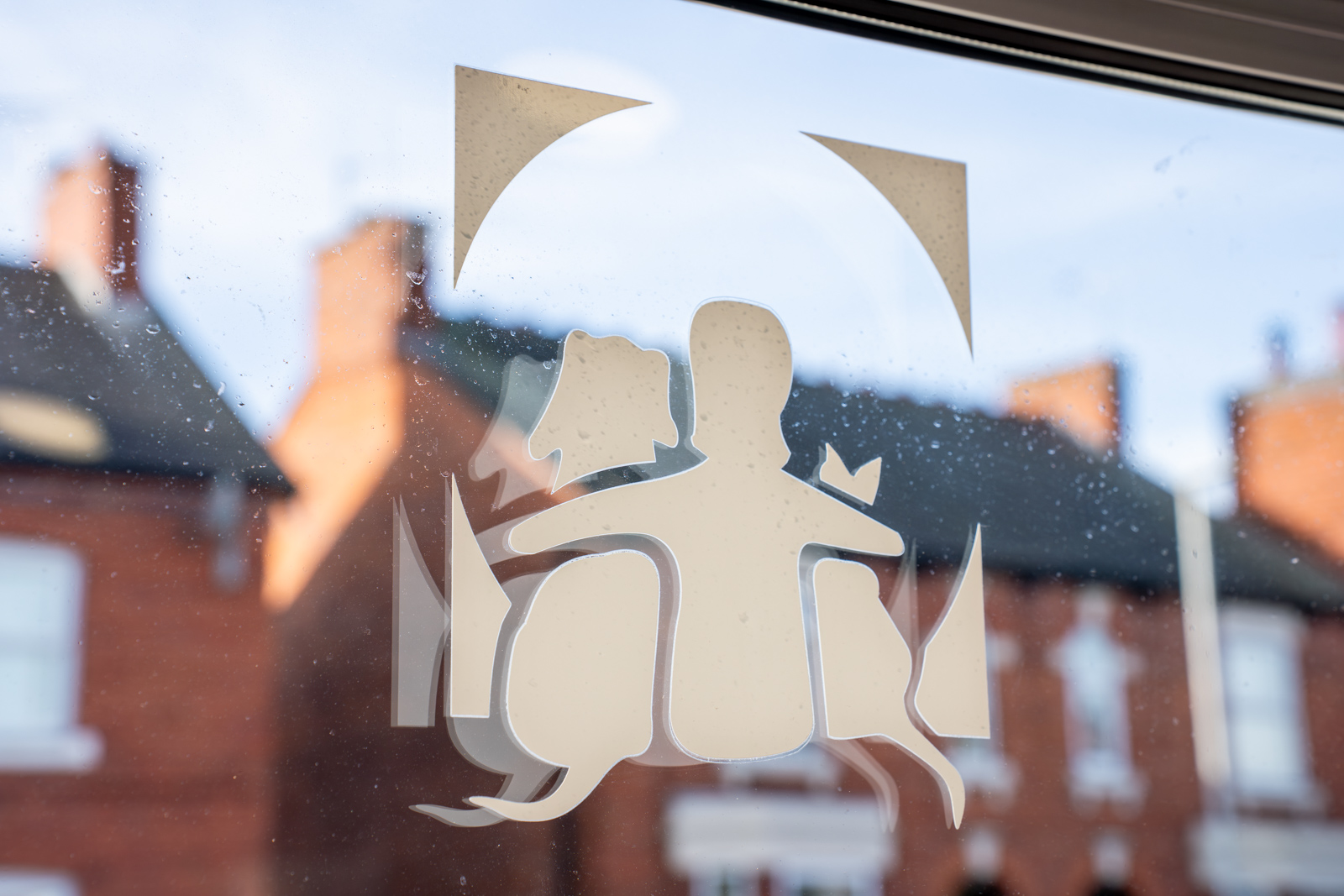 A frosted window decal depicting a stylized person and a dog sitting together. The window overlooks a street with red-brick houses and chimneys in the background. The sky is partly cloudy.
