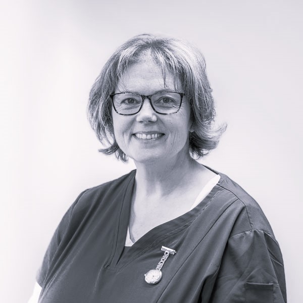 Smiling person with glasses, wearing a dark scrub top and a pin, stands against a plain background.